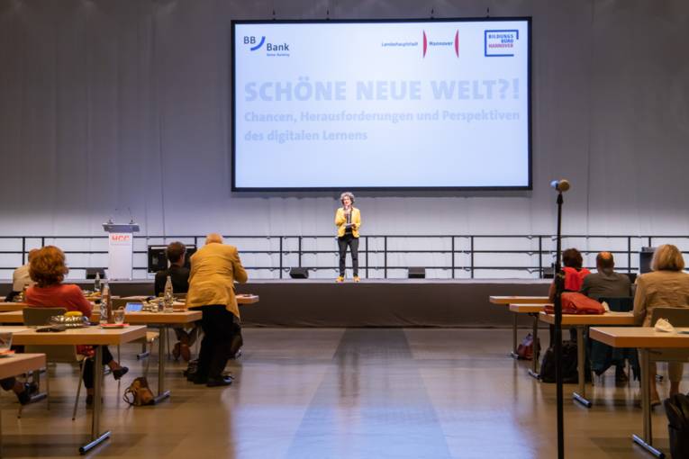 Eine Frau steht in einem Konferenzraum auf der Bühne. Im Vordergrund sitzen Zuhörende. Thema ist "Schöne neue Welt?! Chancen, Herausforderungen und Perspektiven des digitalen Lernens".